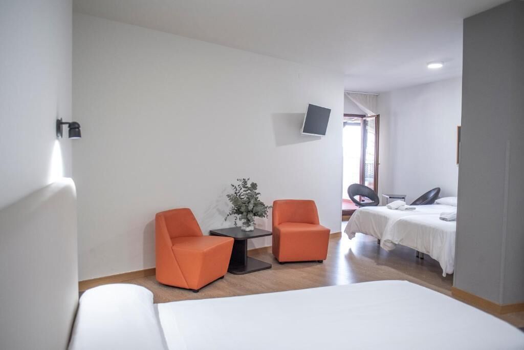 Ein Hotelzimmer mit einem Bett, zwei orangefarbenen Sesseln, einem kleinen Tisch mit einer Pflanze, einem an der Wand montierten Fernseher und einem Fenster mit Vorhängen.