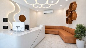Et receptionsområde med en buet, hvid receptionistskranke, en moderne loftlampe, en vægmonteret aircondition og et hjørnesiddeområde med en potteplante.
