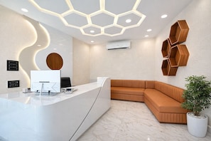 A reception area with a curved white reception desk, a modern ceiling light fixture, a wall-mounted air conditioning unit, and a corner seating area with a potted plant. - Planet Eco Stays (Ujjain)