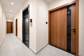 Un passadís amb una porta de fusta, un ascensor negre i un interruptor a la paret.