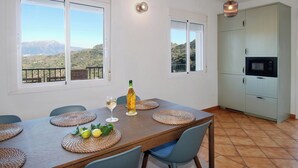 Zona de menjador amb taula preparada per a dues persones, dues finestres amb vistes a la muntanya, cuina amb armaris i llum penjant.