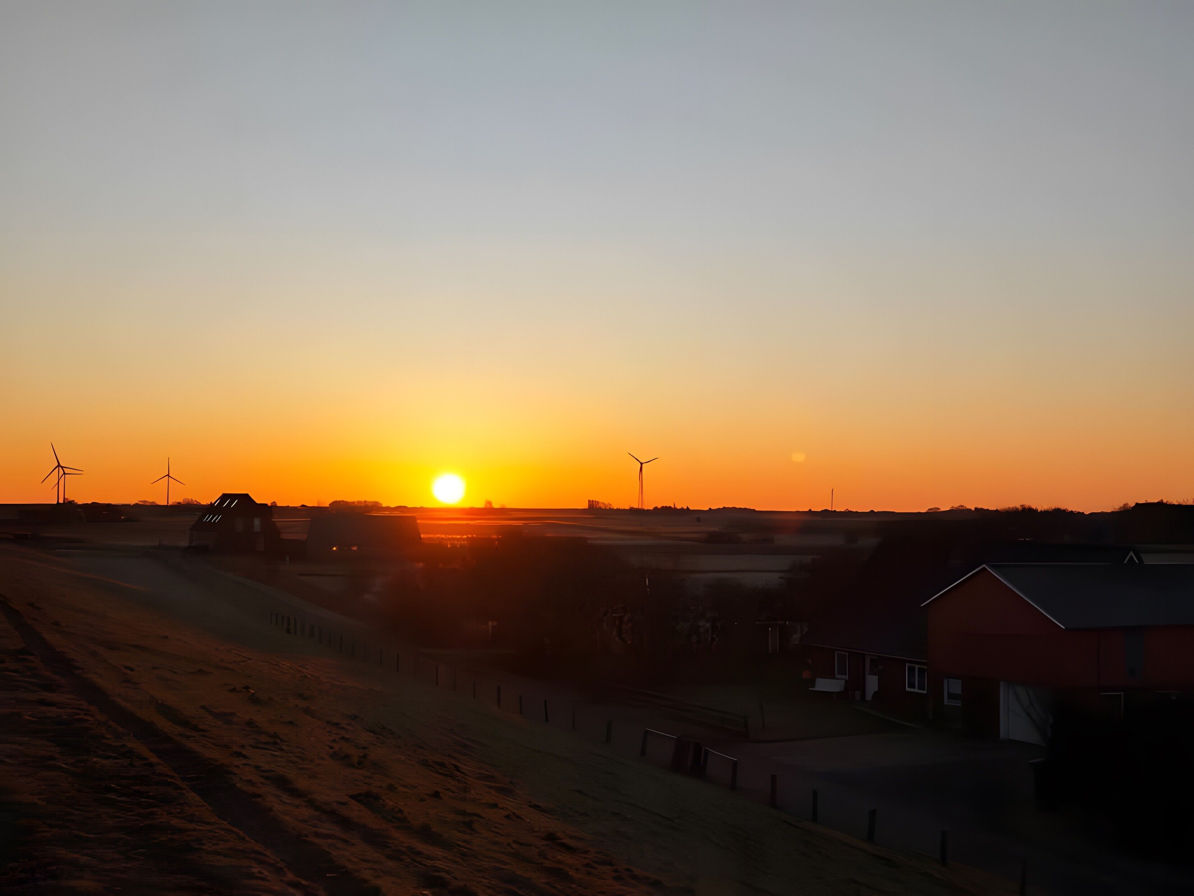 A sunset over a rural landscape with wind turbines and farm buildings.