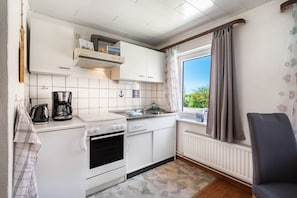 A compact kitchen with white cabinets, a stove, a sink, and a window with curtains. - Apartment 'Ferienhof Knudsen, Seepferdchen' with Shared Terrace, Shared Garden and Wi-Fi (Pellworm)