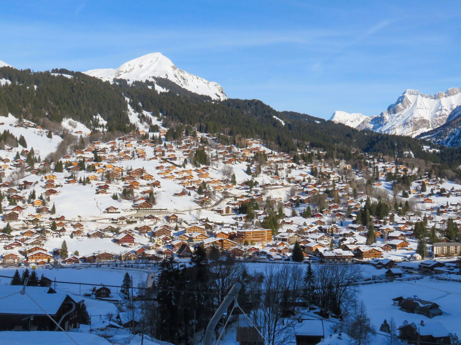 Ein schneebedecktes Dorf mit traditionellen Holzhäusern, einer Seilbahn und Bergen im Hintergrund.