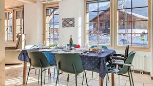 Un comedor con una mesa dispuesta para cuatro personas, ventanas amplias con vista a una montaña nevada y edificios de madera, y un techo con vigas de madera.
