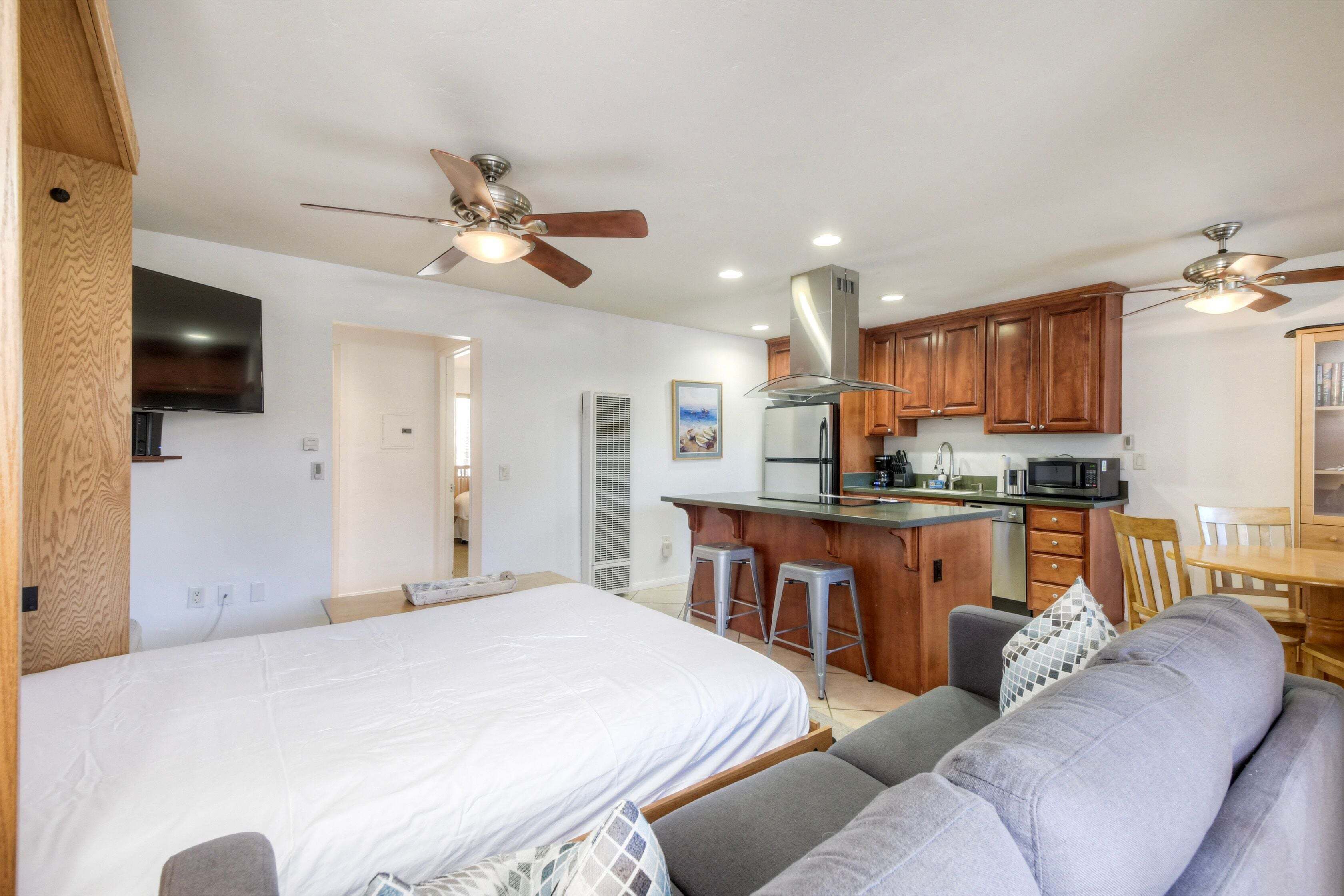 A bedroom with a bed, a gray sofa, a wooden dining table, and a kitchen area with a bar.
