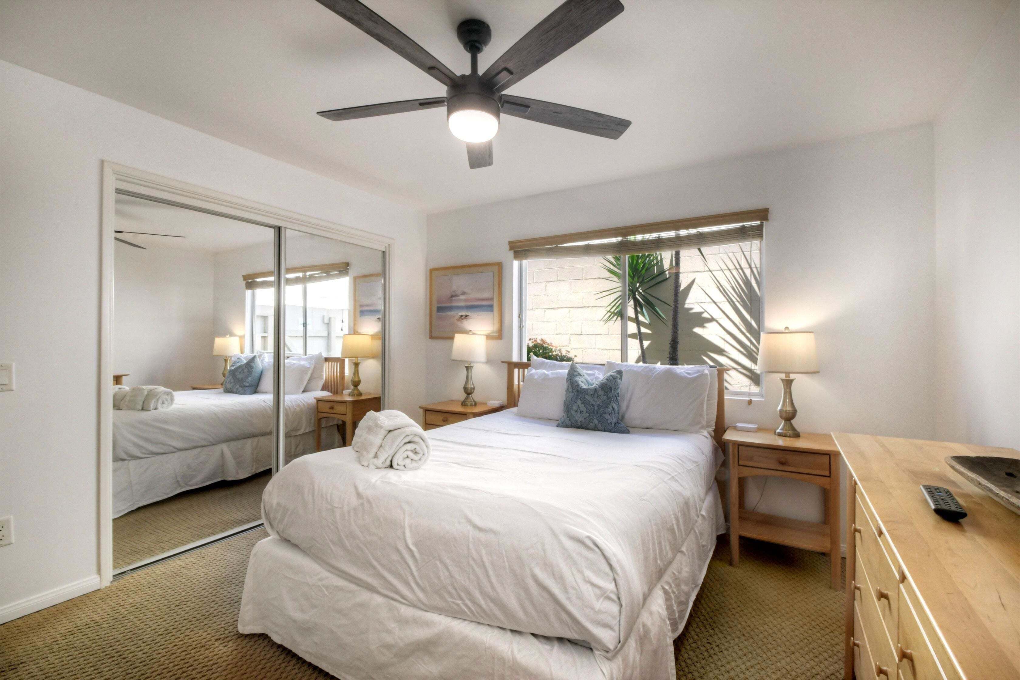 Habitación de hotel con cama, ventilador de techo, ventana con persianas, mesita de noche con lámpara y cómoda de madera.