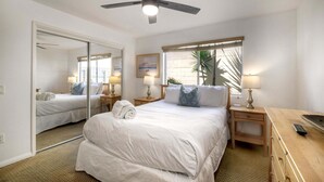 A hotel room with a bed, a ceiling fan, a window with blinds, a nightstand with a lamp, and a wooden dresser. - 90 Walk Score l Private Patio | Steps to Beach & Dining | Parking (Carlsbad)