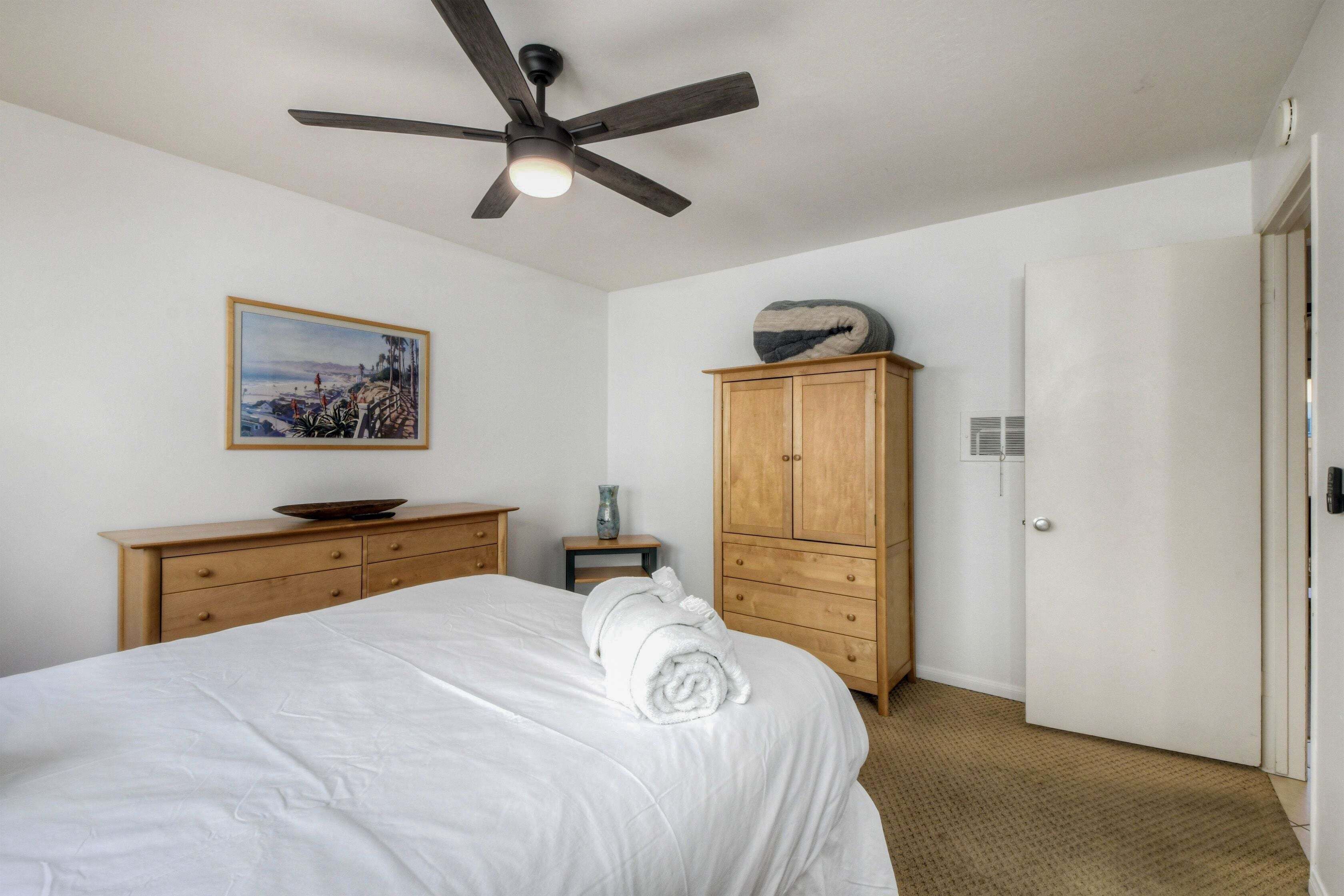 Ein Schlafzimmer mit einem Bett, einem Holzschrank, einem Deckenventilator, einem gerahmten Bild an der Wand und einer Tür.