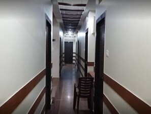 A long, narrow hallway with a wooden chair in the center, white walls, and a dark door at the end. - Hotel Balaji International Araria (Forbesganj)