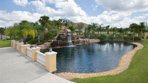 A swimming pool with a waterfall feature, surrounded by palm trees and a paved walkway. - Lovely House for 10 People at Paradise (Four Corners)