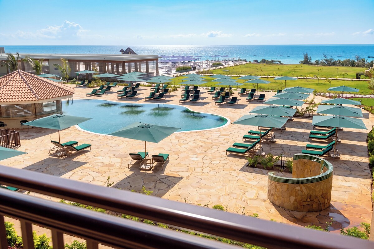 A pool area with lounge chairs and umbrellas, a stone deck, and a view of the ocean.