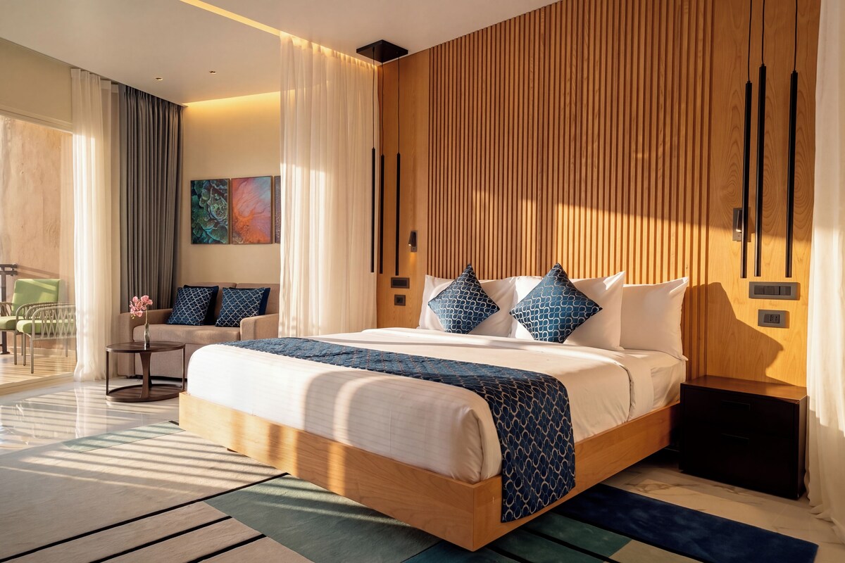 A modern hotel room with a large bed, a seating area with a sofa and chairs, and a view of the outdoors.