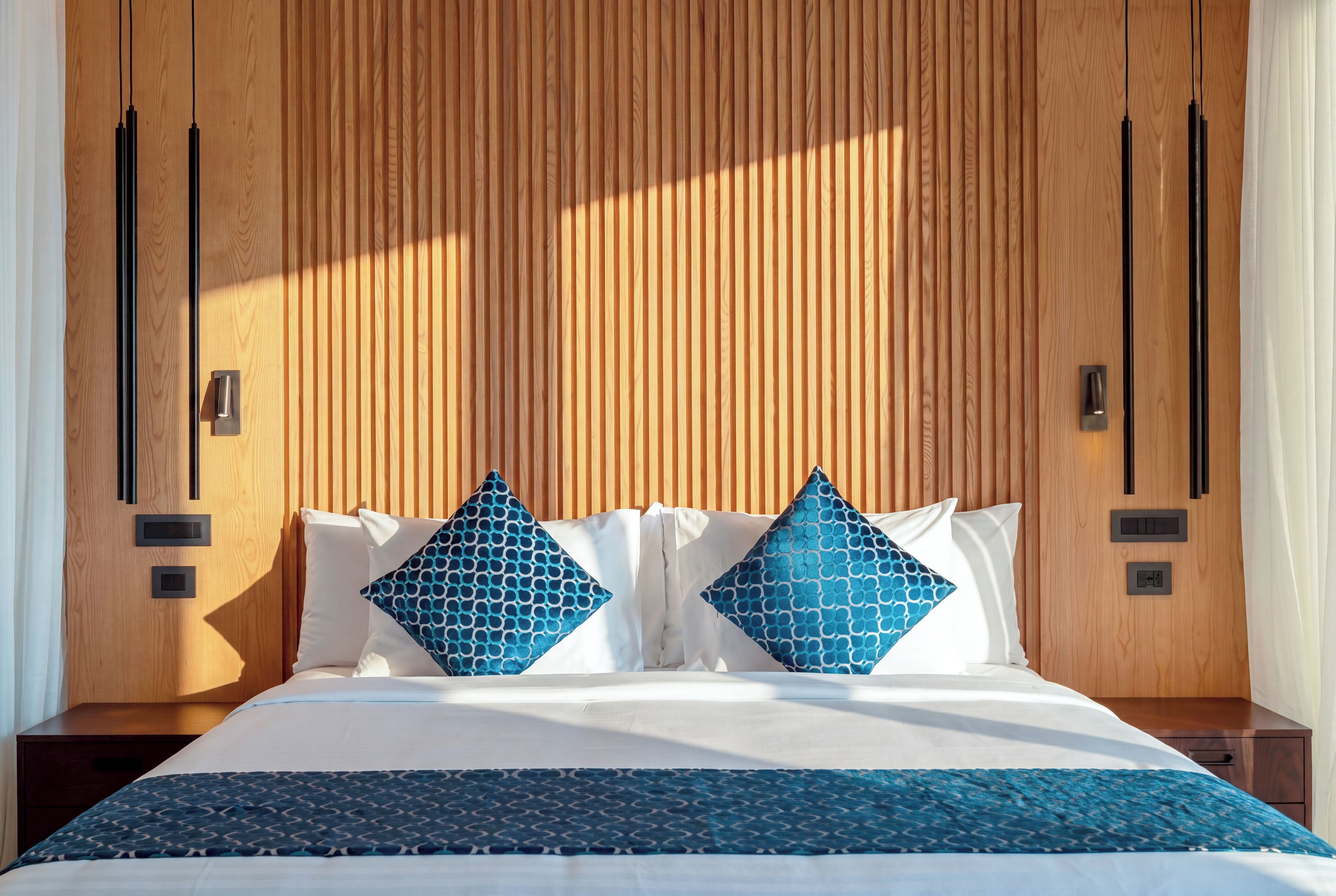Habitación de hotel con cabecera de madera, cama con sábanas blancas, dos almohadas estampadas en azul y una manta estampada en azul.