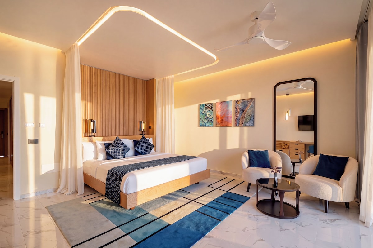 A modern hotel room with a large bed, a seating area with a coffee table, and a large mirror.