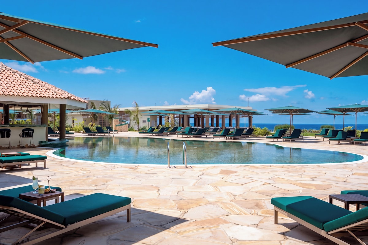 A pool area with lounge chairs and umbrellas, a clear sky, and a view of the ocean.