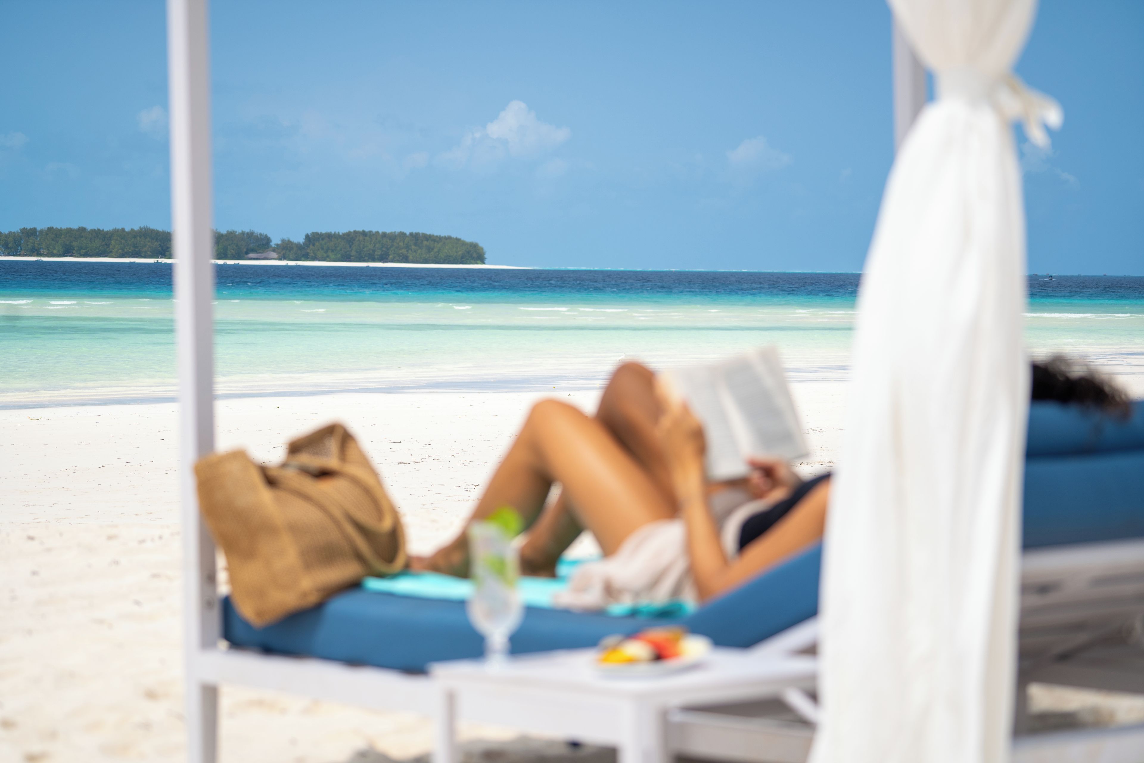 A beachside lounge chair with a person reclining, a drink, and a book, overlooking a clear blue sea and a distant island.