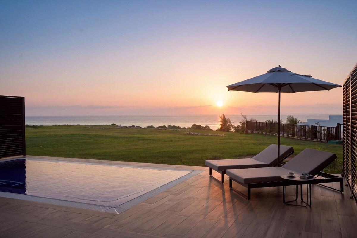 A poolside area with lounge chairs and an umbrella, overlooking a grassy field and the ocean at sunset.