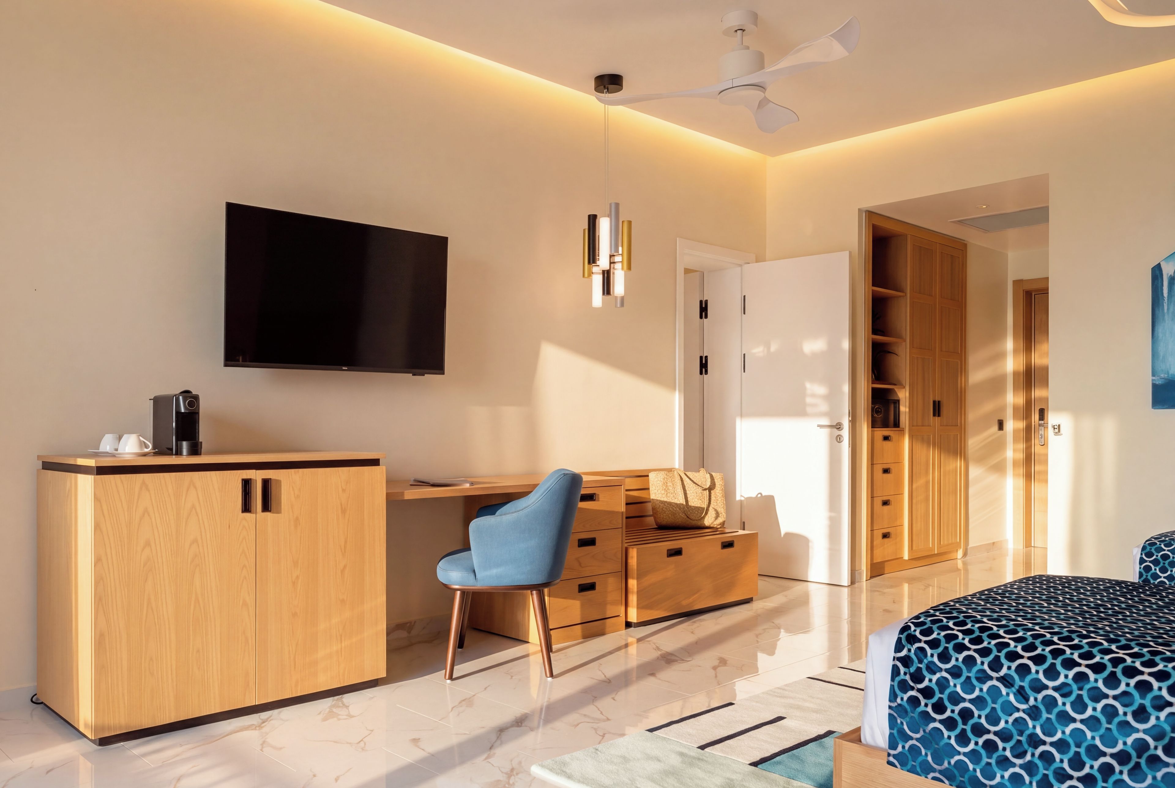 Une chambre d’hôtel moderne dotée d’une télévision à écran plat, de meubles en bois, d’un bureau, d’une chaise bleue, d’une armoire et d’un lit avec une literie à motifs.