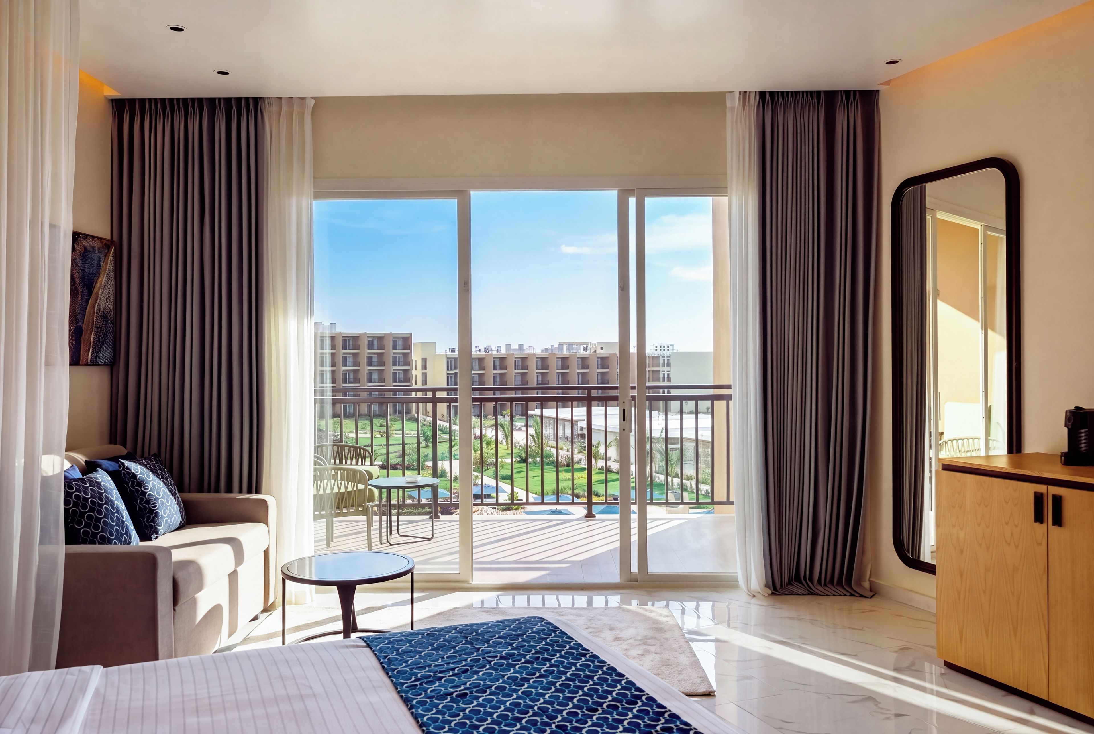 A hotel room with a bed, a sofa, a small table, a balcony with a view of the city, and a large mirror.