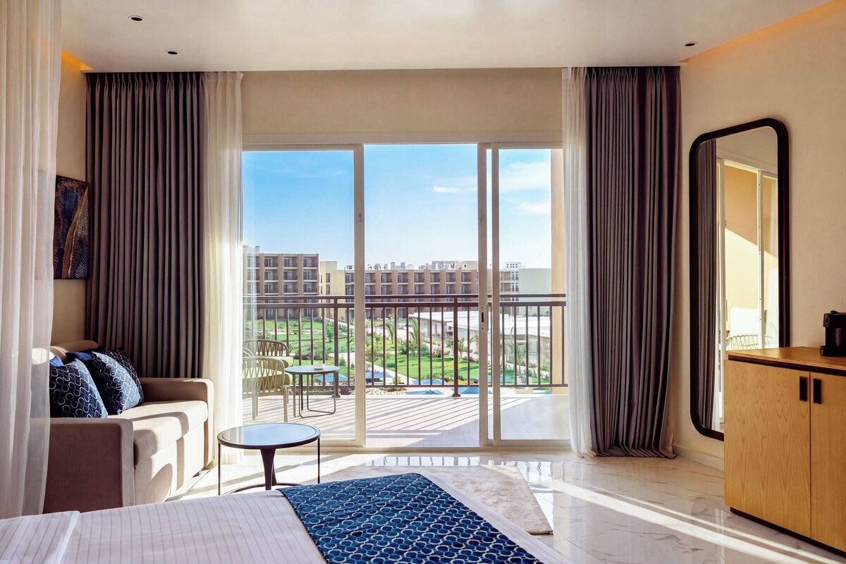 A hotel room with a bed, a seating area, a small table, a balcony with a view of the city, and a large mirror.