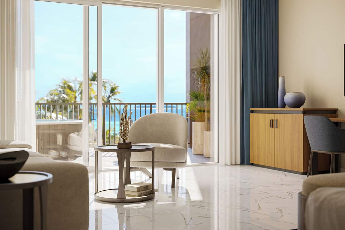 Habitación de hotel moderna con un amplio ventanal corredero que da a un balcón con vistas al mar. Hay una mesa redonda con un jarrón y libros, un sofá y un mueble de madera con adornos.