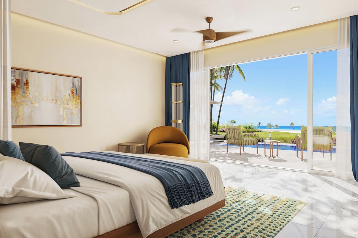 Habitación de hotel con cama, ventilador de techo, ventana grande con vista a la playa y una pequeña mesa con sillas.
