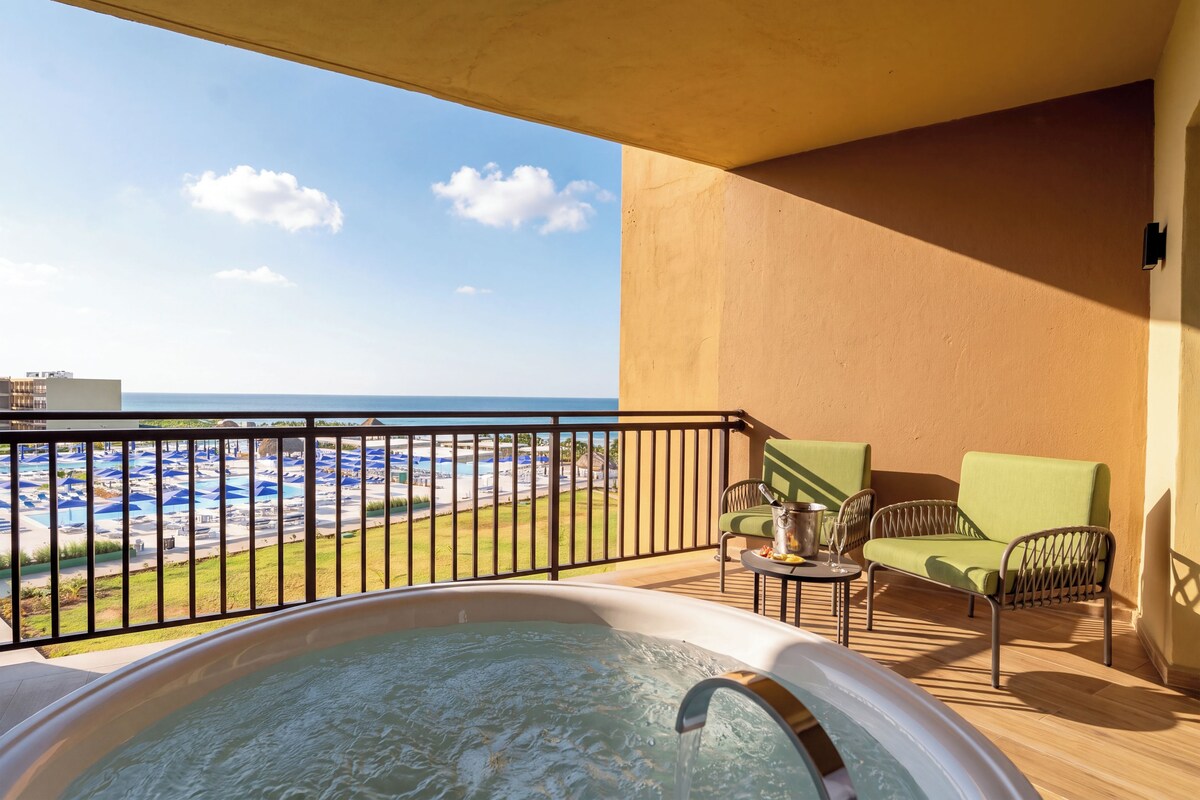 A balcony with a hot tub, a table with a bottle and glasses, and a view of a pool and beach.