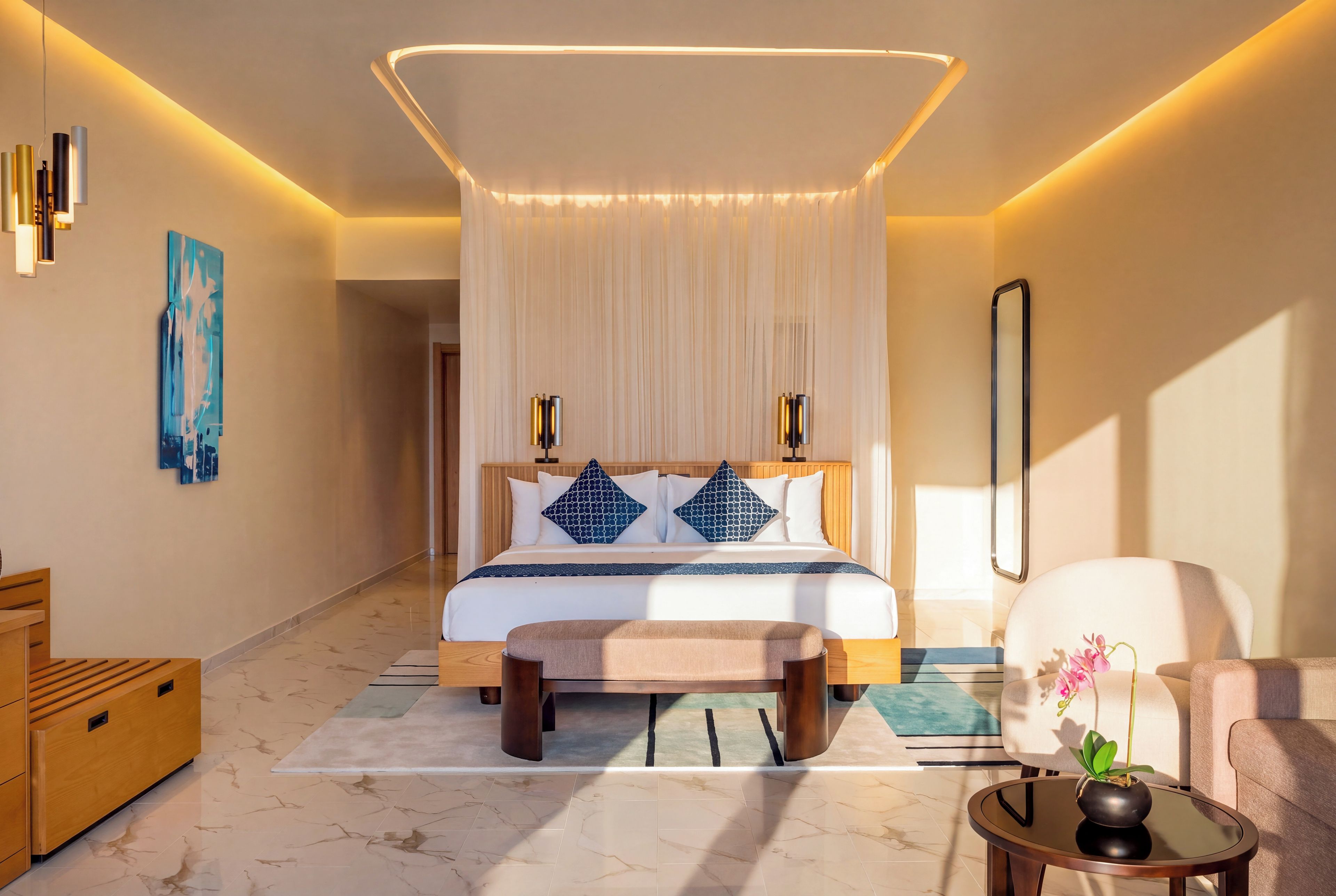 A modern hotel room with a large bed, bedside lamps, a bench, a small table with a plant, and a painting on the wall.