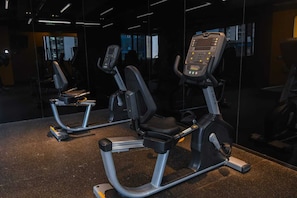Dos bicicletas estáticas en un gimnasio con un muro espejado y una ventana al fondo.