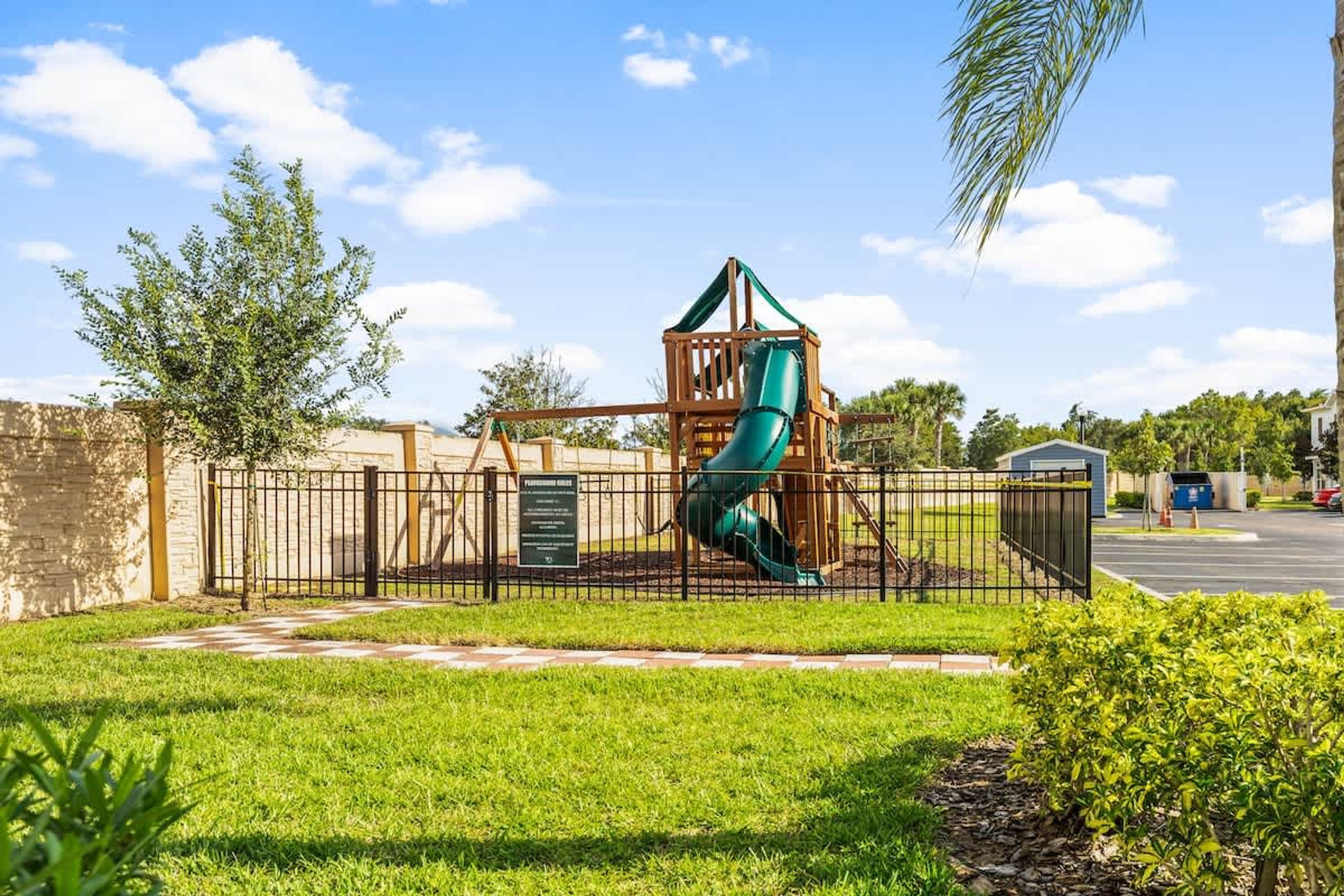 A playground with a slide and climbing structure, surrounded by a fence and greenery.