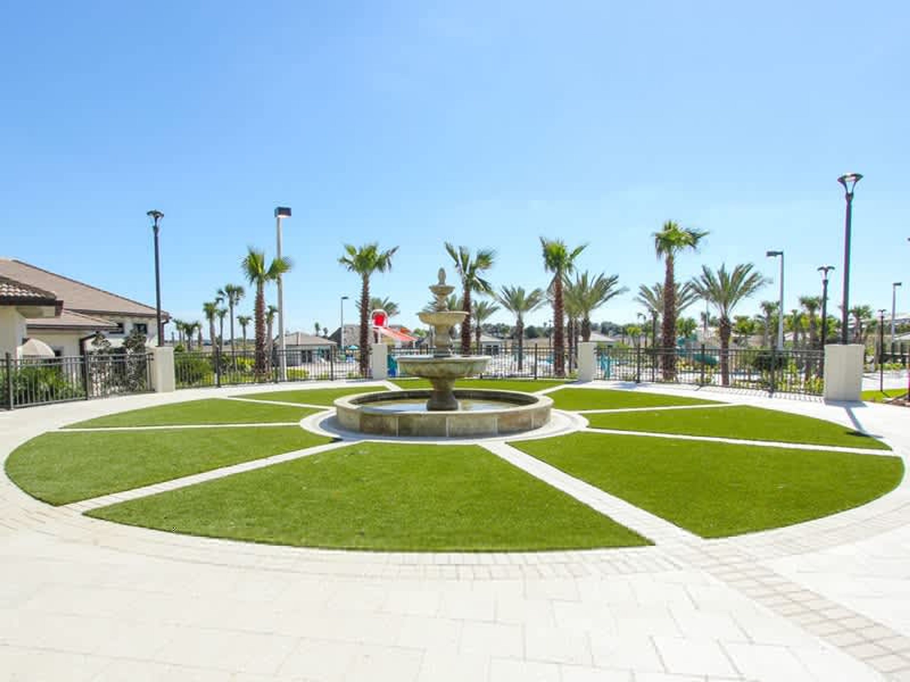 Une place publique bien entretenue, avec une fontaine centrale, entourée de pelouses soigneusement taillées et de palmiers.