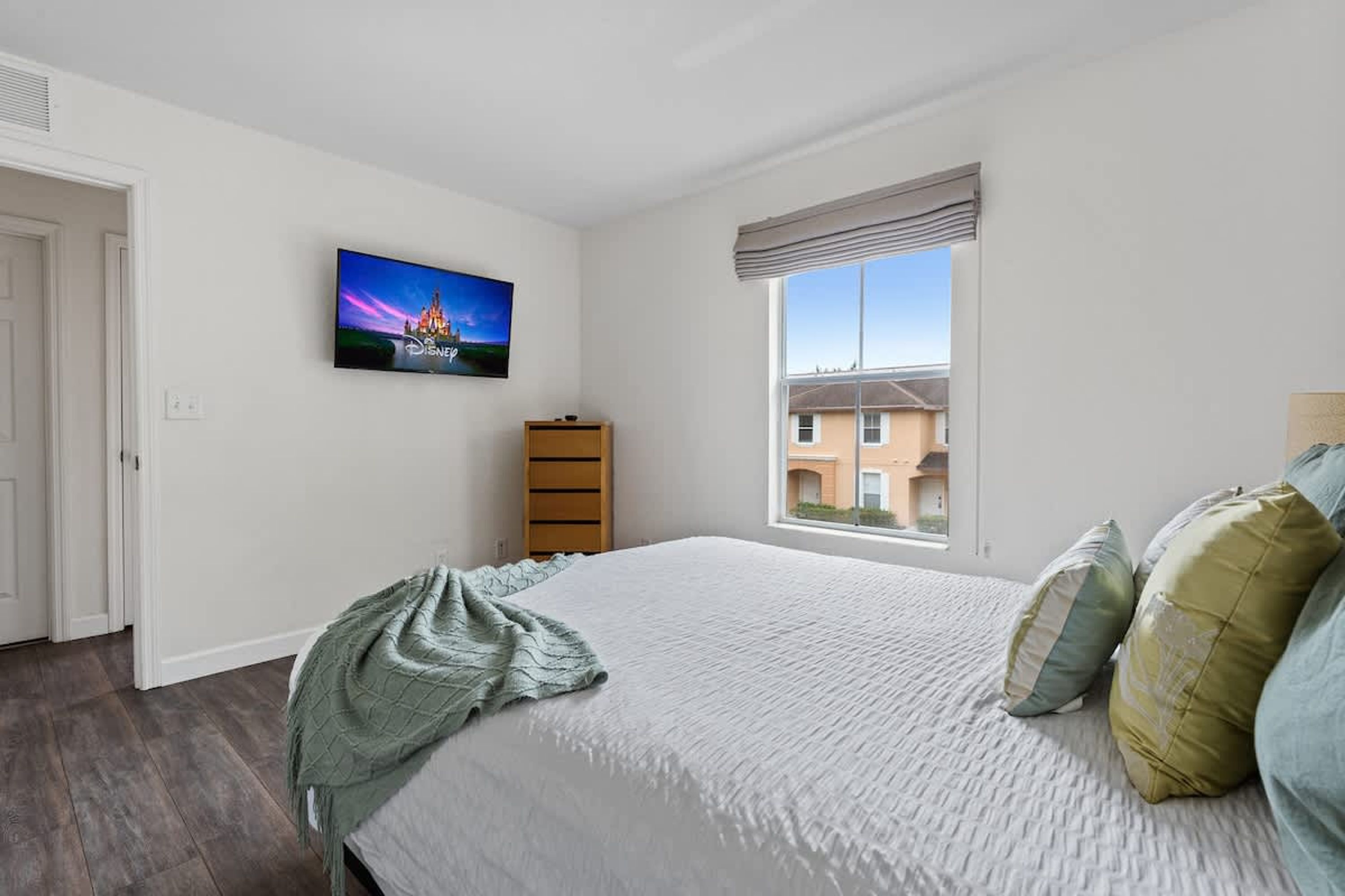A bedroom with a bed, a television on the wall, a window with a view of buildings, and a door.