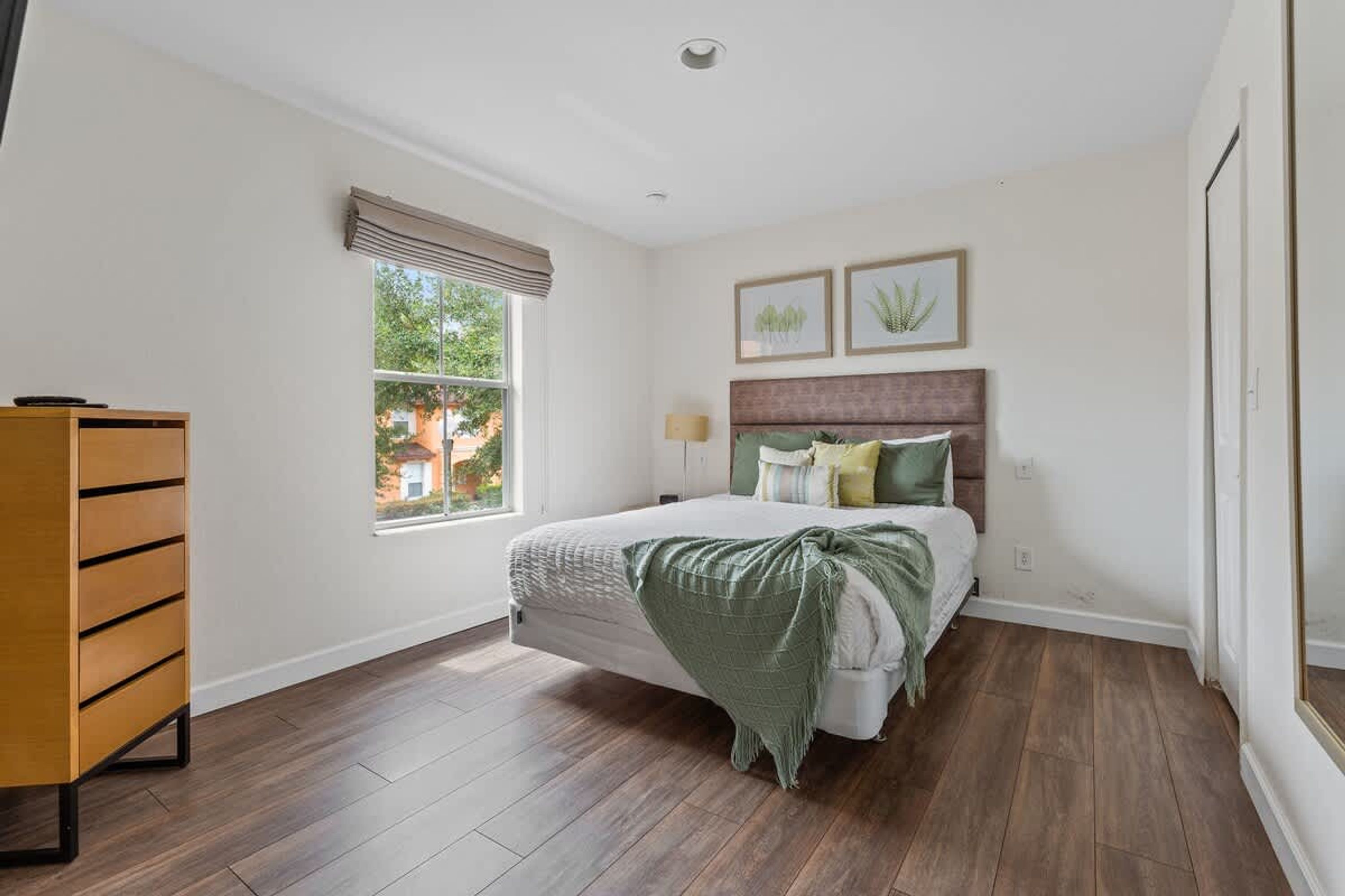 Een slaapkamer met een bed, een kaptafel, een raam met jaloezieën en twee ingelijste foto's aan de muur.