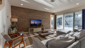 A modern living room with a large flat-screen TV, grey sofas, wooden chairs, and a checkered floor. - Amazing House at Bella Vida 5BR (Kissimmee)