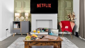 En stue med et fladskærms-tv der viser 'NETFLIX', en pejs, et sofabord med frugt og drikkevarer, en bærbar computer og en hyggelig lænestol.