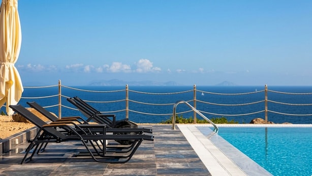 Area piscina con sedie a sdraio e un ombrellone grande, con vista su un mare azzurro e montagne in lontananza.