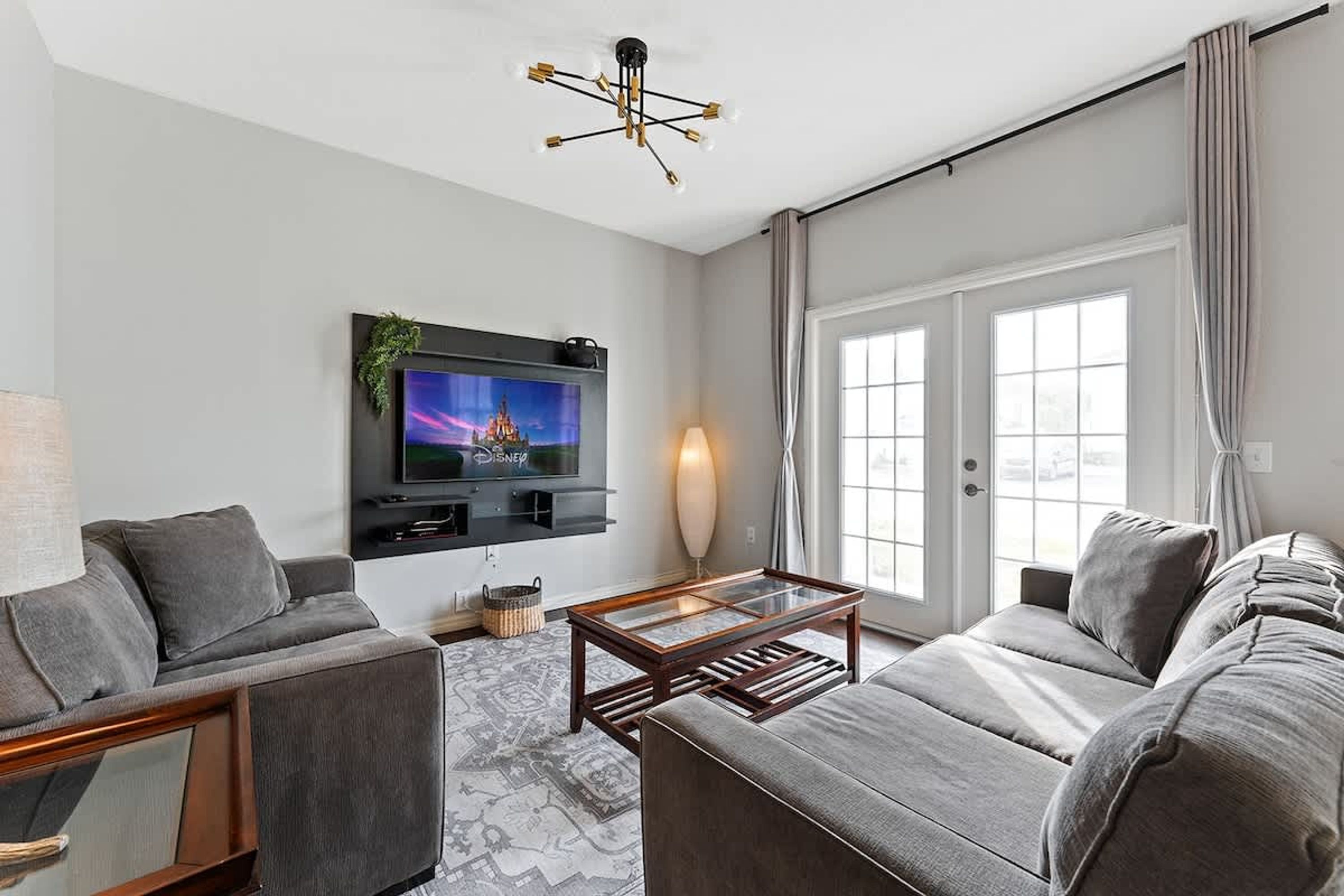 Ein modernes Wohnzimmer mit grauem Sofa, einem Couchtisch, einem an der Wand befestigten Fernseher und einem großen Fenster mit Vorhängen.