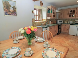 A dining area with a wooden table set for four, a vase of flowers, and a kitchen in the background.