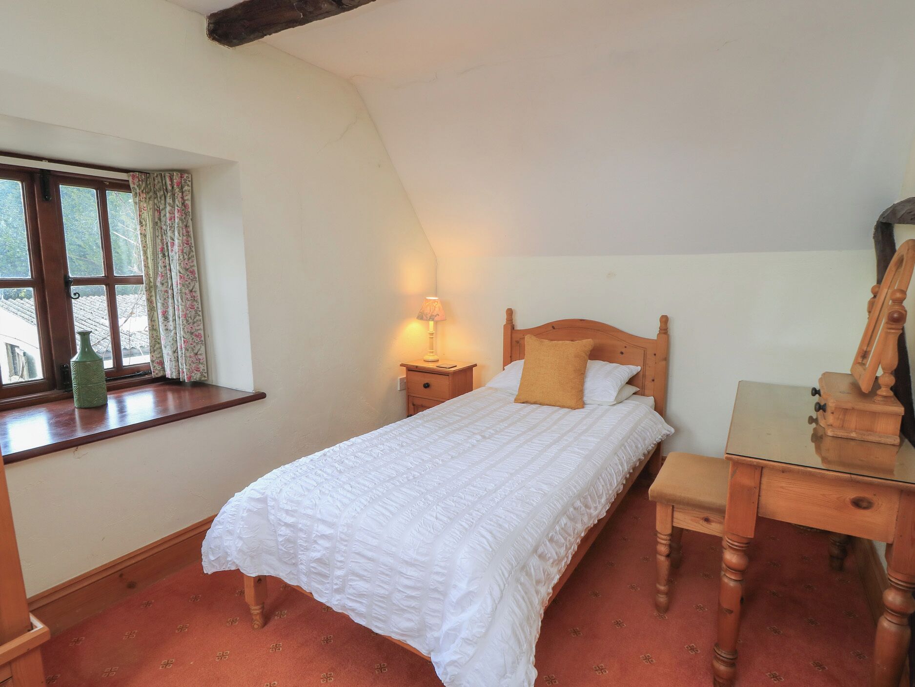 A bedroom with a bed, a nightstand, a lamp, a window with curtains, and a wooden desk.