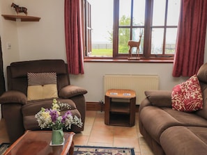 A living room with a brown sofa, a brown armchair, a small table with a vase of flowers, and a window with a view of greenery.