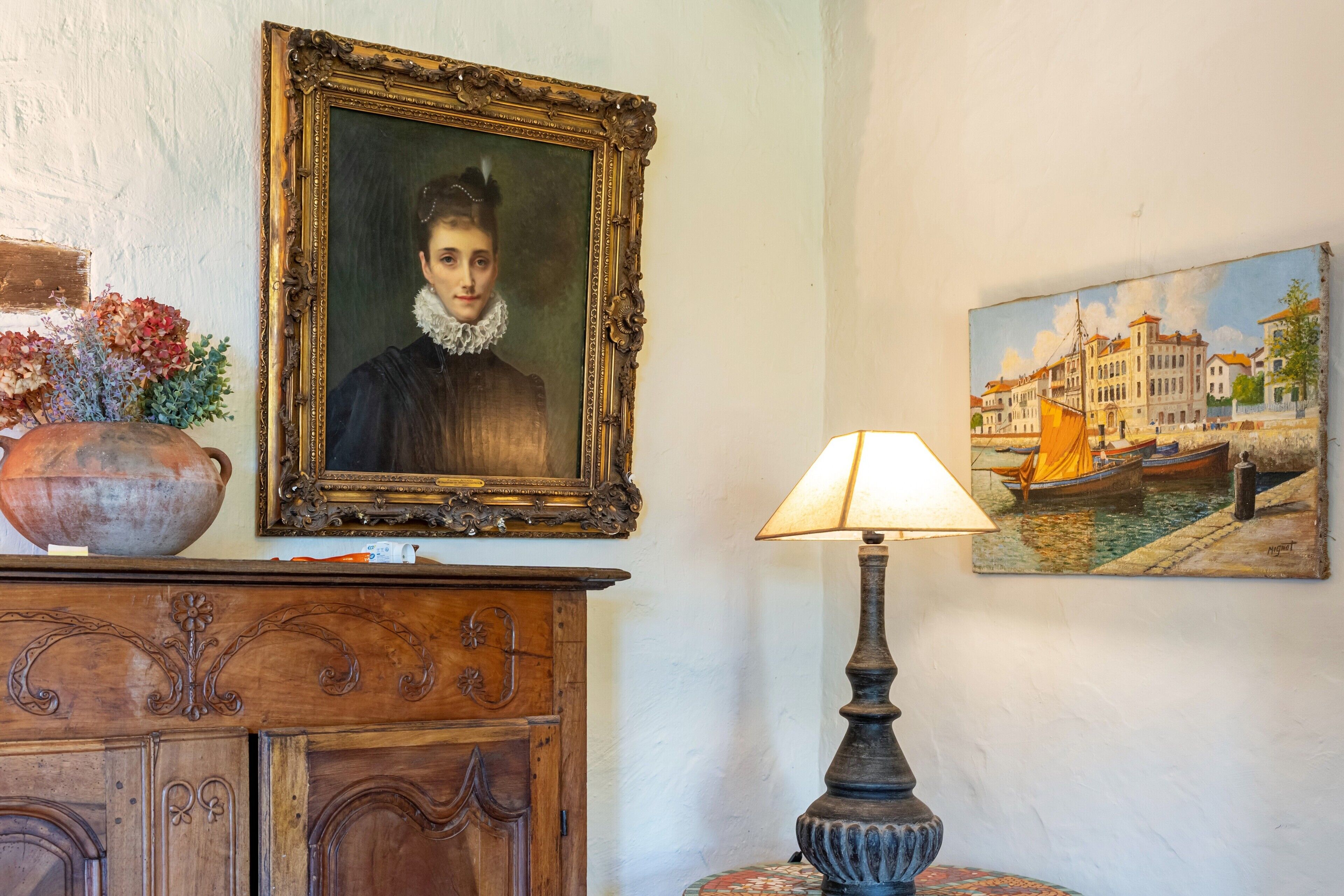 A room with a wooden cabinet, a framed portrait, a lamp, and a painting of a canal scene.