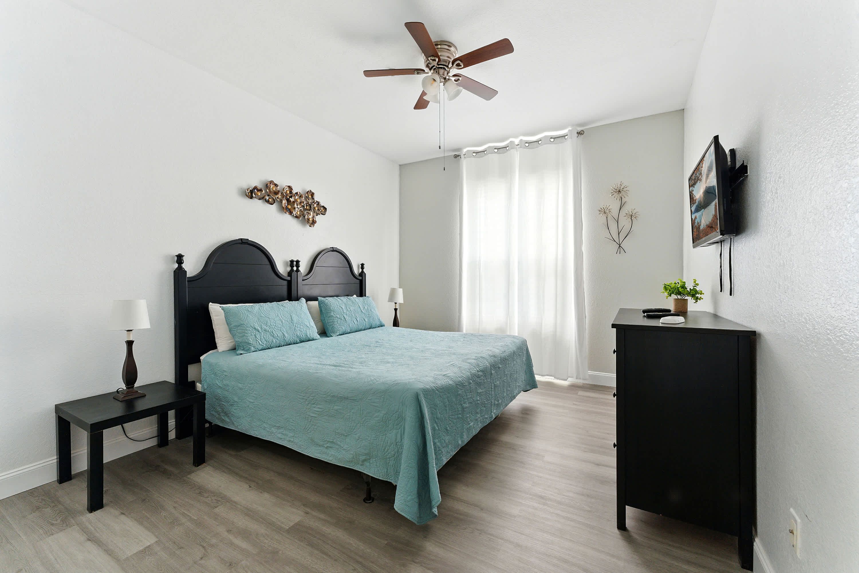A bedroom with a bed, nightstand, dresser, ceiling fan, and a wall-mounted TV.