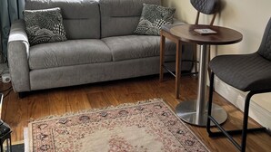 A cozy living room with a grey sofa, patterned cushions, a small round table, a dark armchair, a patterned rug, and a large window with curtains.