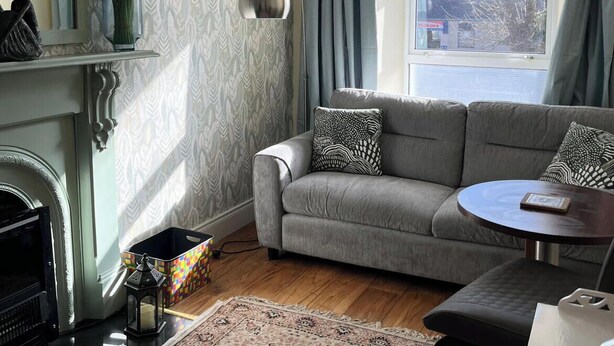 A living room with a grey sofa, a small round table, a fireplace, a mirror, and a window with curtains.