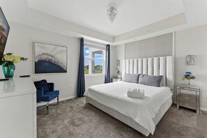 Een moderne slaapkamer met een groot bed, een bureau en een stoel. Er is een raam met gordijnen, een schilderij aan de muur en een kroonluchter.