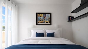 Hotellihuone, jossa on sänky, valkoiset seinät, kehystetty taulu ja ikkuna, jossa on verhot.