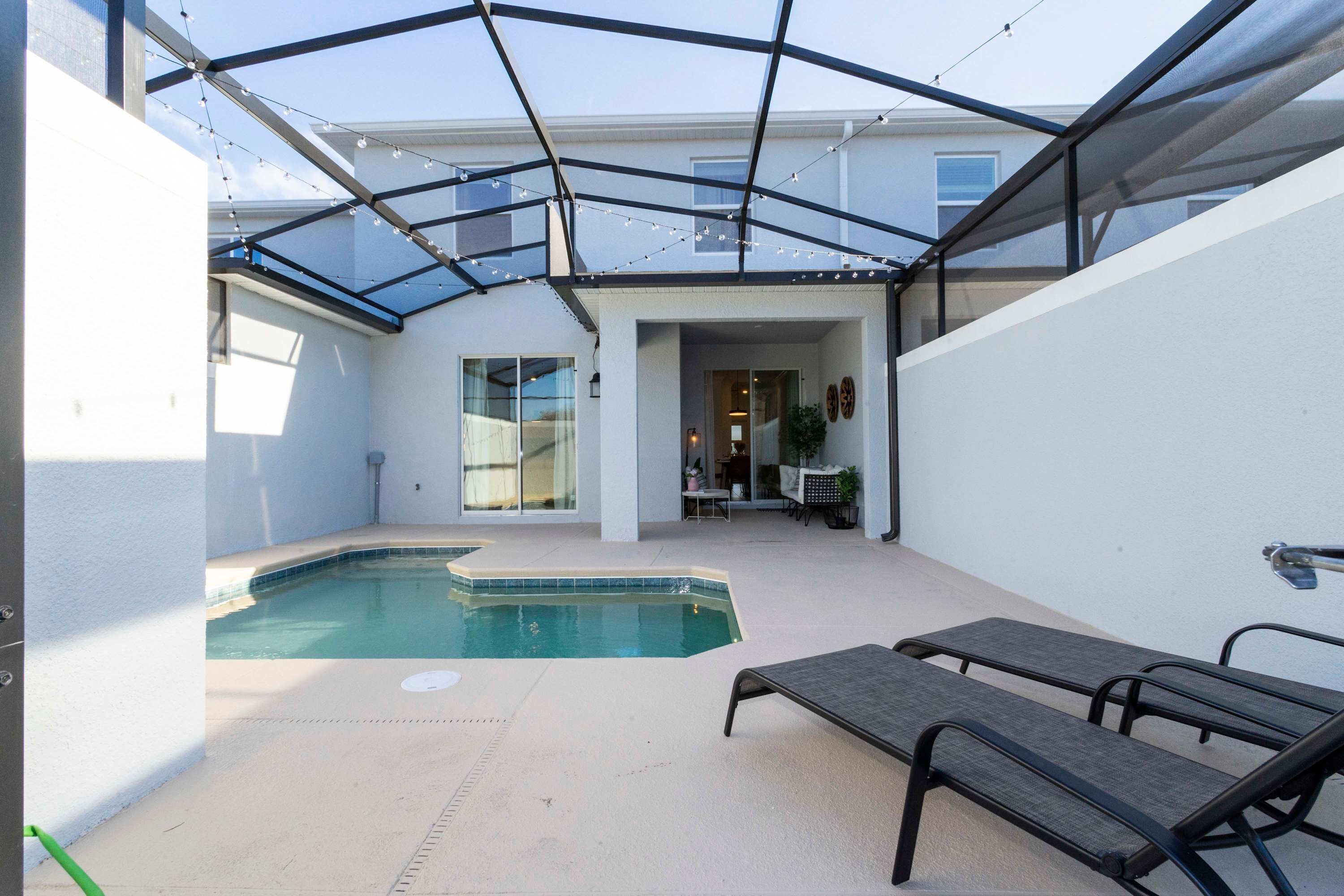 A modern outdoor pool area with a glass enclosure, lounge chairs, and a patio with a table and chairs.