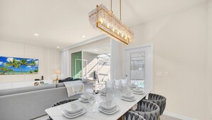 A dining area with a white table set for six, grey upholstered chairs, and a crystal chandelier. - Stylish 5BR Home with Mickey & Cars Rooms (Four Corners)