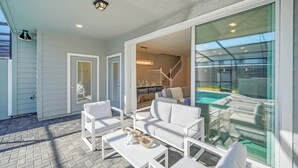 A spacious outdoor patio with white furniture, a glass-enclosed dining area, and a view of a pool. - Stylish 5BR Home with Mickey & Cars Rooms (Four Corners)
